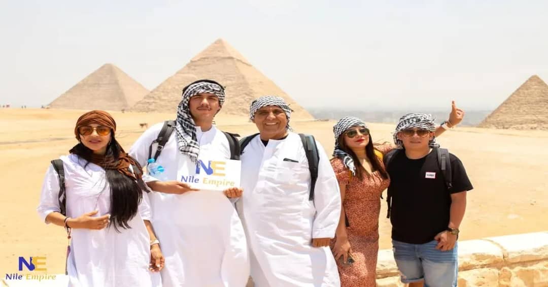 Cinco viajeros con galabiyas blancas y pañuelos posando con un cartel de Nile Empire ante las pirámides de Giza