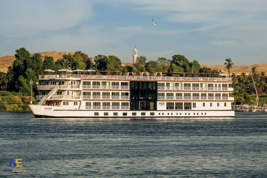 Barco de crucero de cuatro cubiertas navegando por el Nilo junto a una orilla con palmeras, un minarete y colinas desérticas