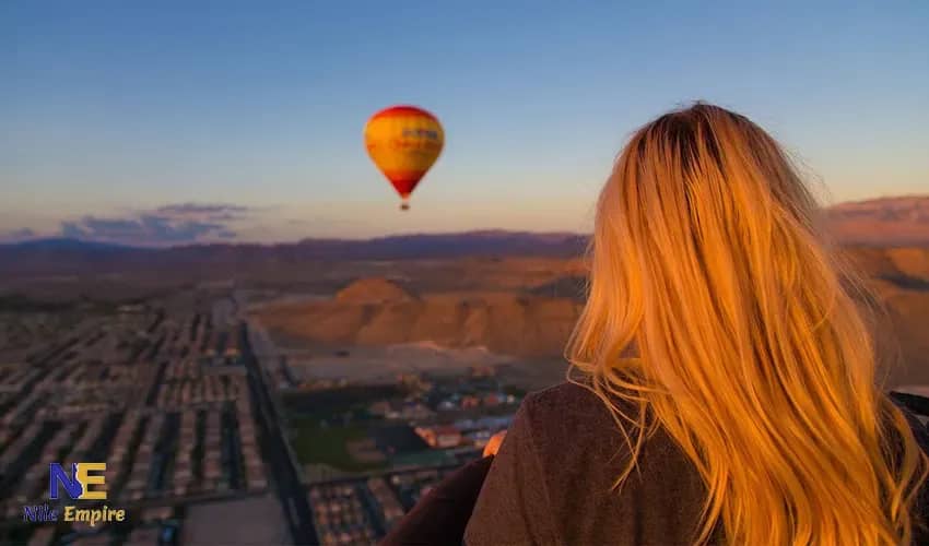 Viajera de espaldas contemplando un globo aerostático rojo y amarillo al amanecer sobre una ciudad y colinas desérticas