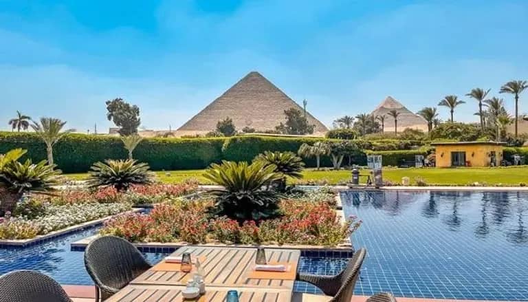 Terraza con mesa junto a una piscina y jardines con palmeras, con dos pirámides de Giza al fondo