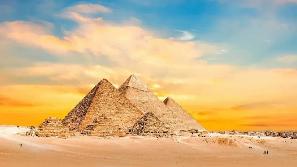 Las pirámides de Giza al atardecer bajo un cielo dorado, con algunos visitantes caminando por la arena del desierto