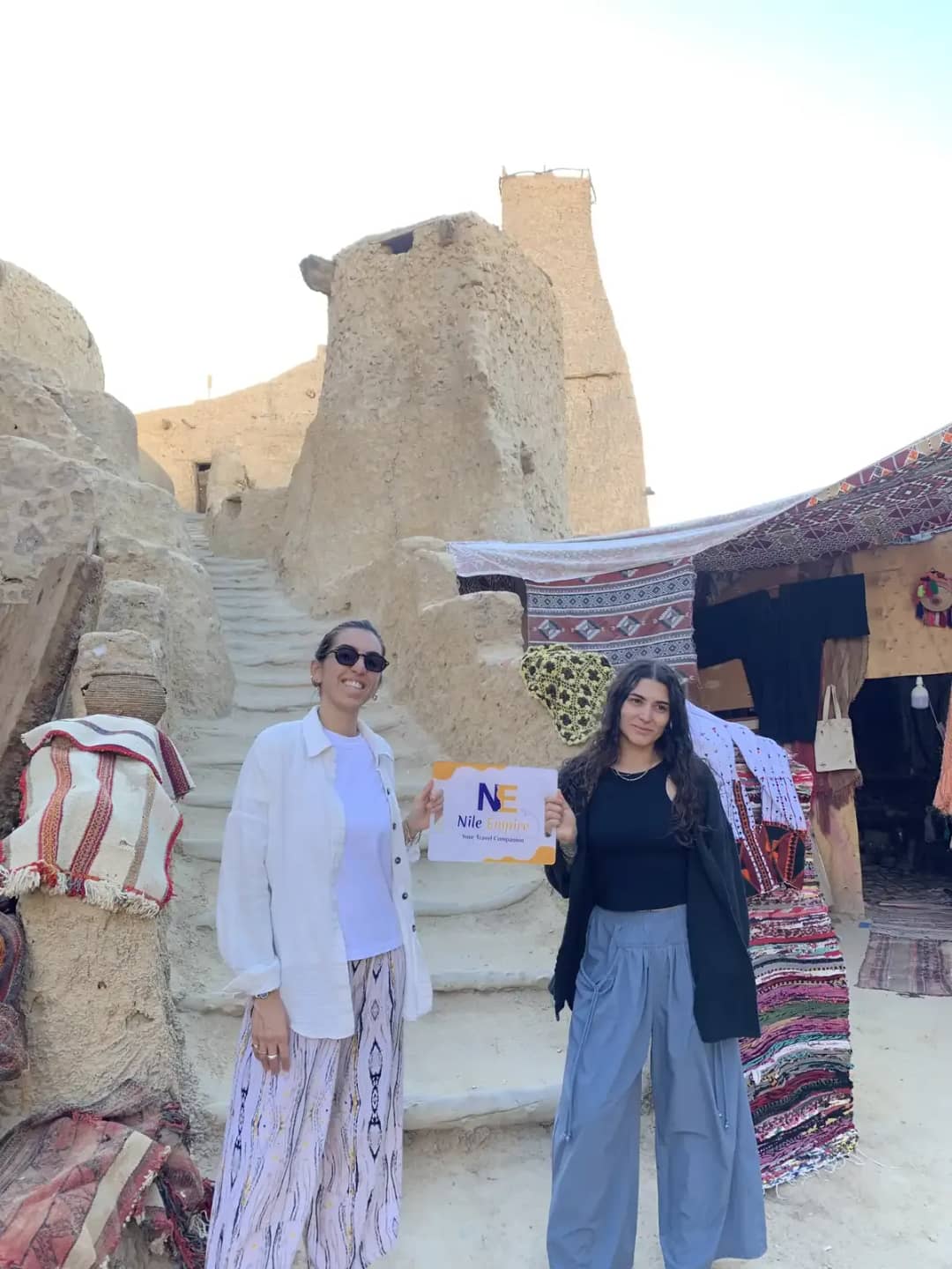 Two travellers holding a tour sign beside a textile stall in a mud-brick desert village