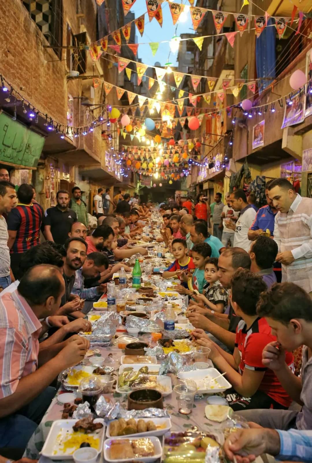 Iftar comunitario en una calle de El Cairo durante el Ramadán: largas mesas con comida, vecinos y niños sentados bajo banderines, globos y guirnaldas de luces